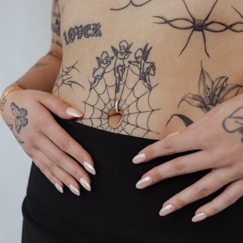 Bauchnabel mit elegantem Titan Clicker und Kristall, umgeben von Tattoos auf hellem Hintergrund