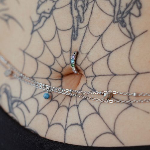 Bauchnabel-Piercing mit Regenbogen-Kristallen auf tätowierter Haut, Nahaufnahme
