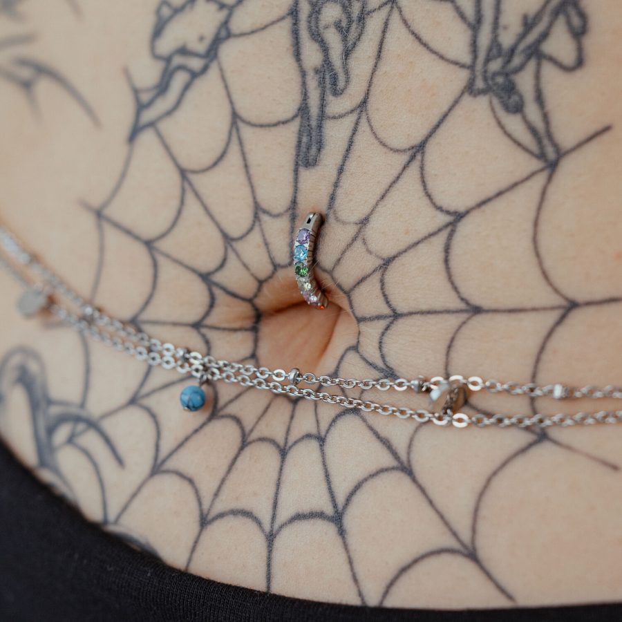 Bauchnabel-Piercing mit Regenbogen-Kristallen auf tätowierter Haut, Nahaufnahme