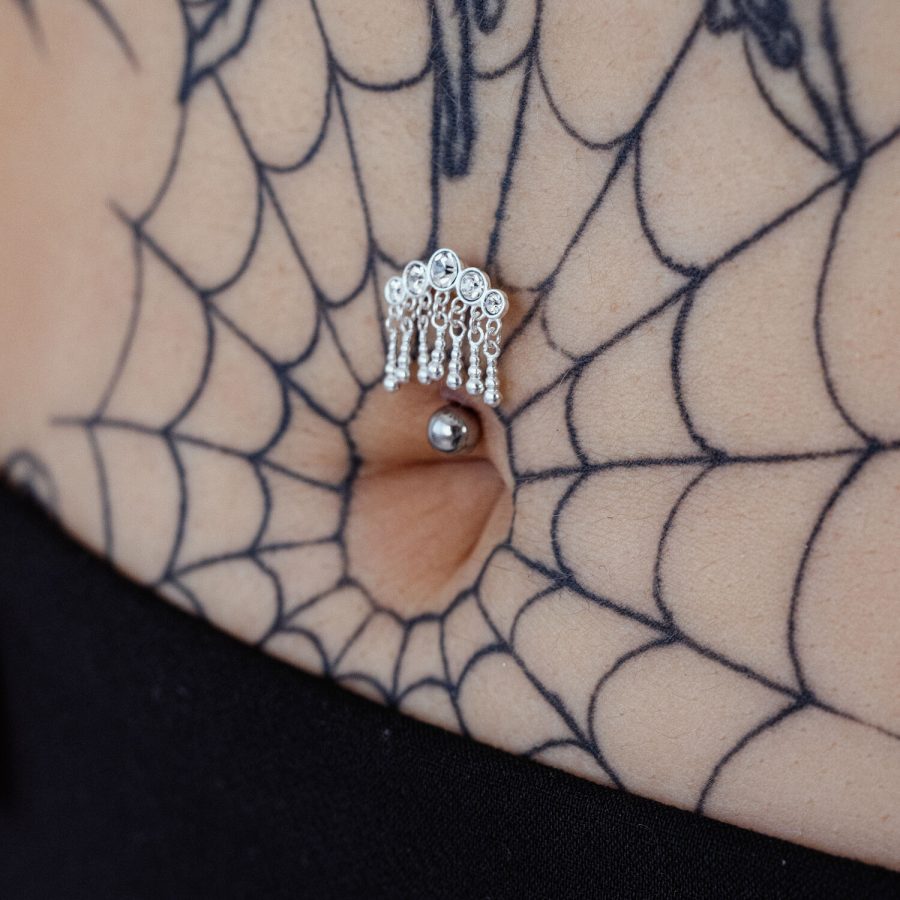 Silbernes Bananabell-Piercing mit fünf hängenden Kristallen in einem Bauchnabel, umgeben von einem Spinnennetz-Tattoo