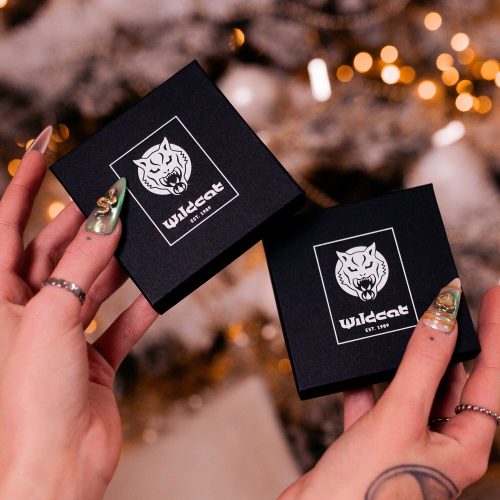 Zwei schwarze Geschenkboxen mit Wildcat-Logo, gehalten von Händen mit goldfarbenen Nägeln