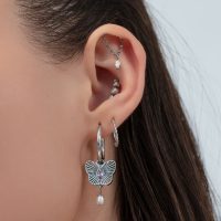 Nahaufnahme eines Ohres mit silbernen Creolen, Schmetterlingsanhänger und mehreren Piercings vor hellem Hintergrund