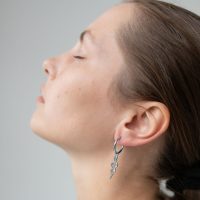 Profil einer Person mit geschlossenen Augen, die silberne Schlangen-Ohrringe trägt, auf grauem Hintergrund.