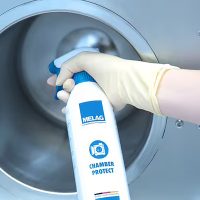 Hand mit gelbem Handschuh hält Sprühflasche Chamber Protect vor rundem Metallkessel