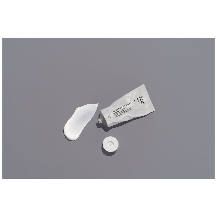 Graue Tube der Nø Cosmetics Intensiv Repair Cream mit abgenommenem Deckel und etwas Creme daneben auf grauem Hintergrund