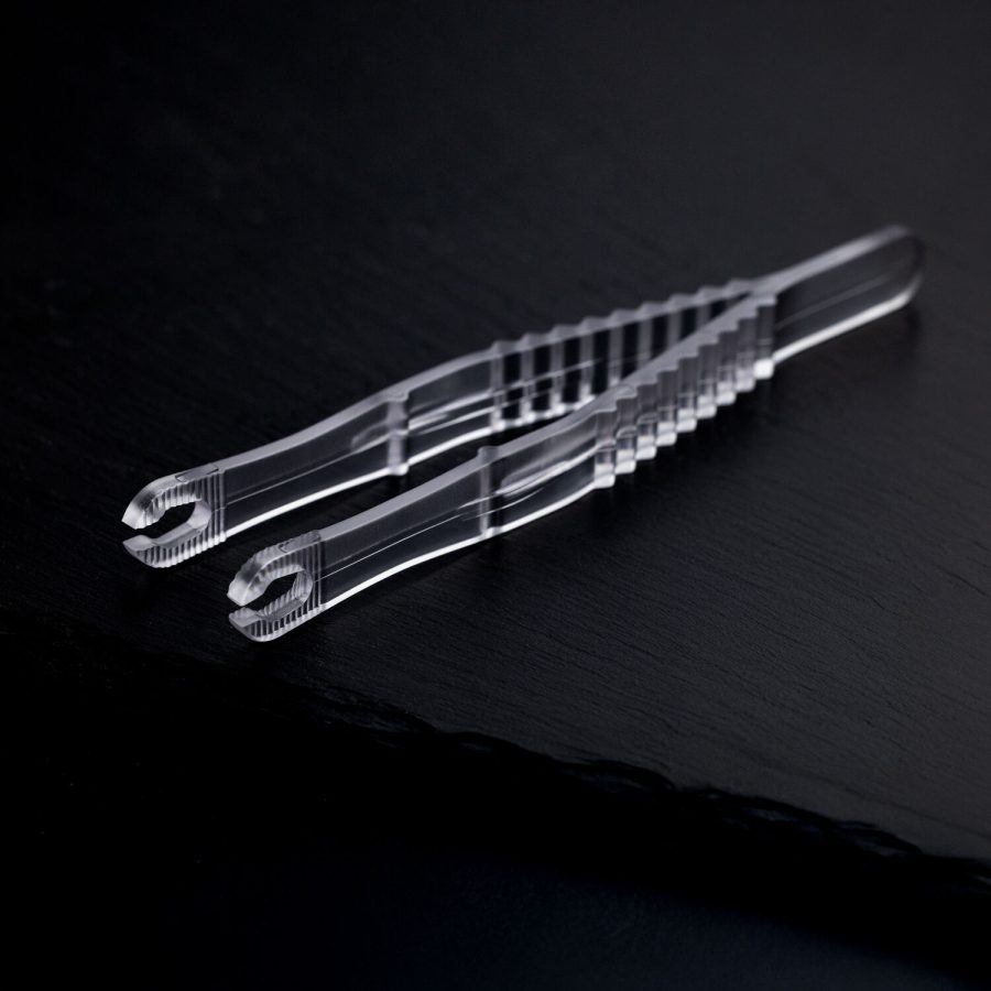 Zwei transparente, sterile Runde Piercingzangen liegen überkreuzt auf einem dunklen Hintergrund.