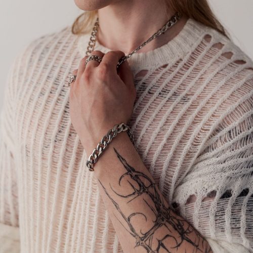 Person mit tätowiertem Arm trägt ein silbernes Chunky Armband und einen gestrickten Pullover