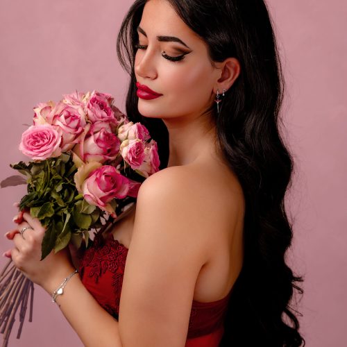 Frau mit langen, dunklen Haaren hält einen Strauß rosa Rosen vor rosa Hintergrund.