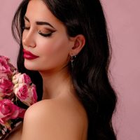 Frau mit langen dunklen Haaren und silbernen Ohrringen mit roten Kristallen, hält rosa Blumen vor rosa Hintergrund