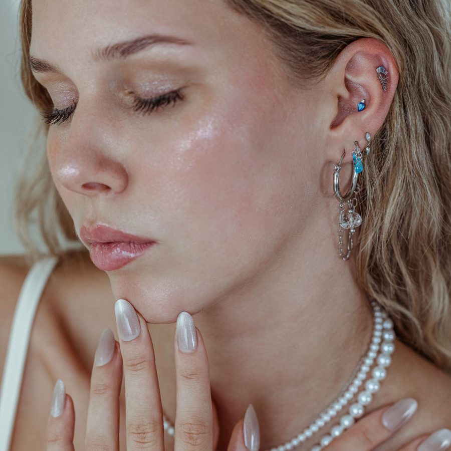 Frau mit mehreren Ohrpiercings, darunter der Mystic Dew Aufsatz in Silber und Gold mit Lavendel- und Türkisfarbenen Kristallen, auf neutralem Hintergrund