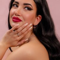 Frau mit langen schwarzen Haaren, roten Lippen und silbernem Ohrring mit rotem Kristall vor rosa Hintergrund
