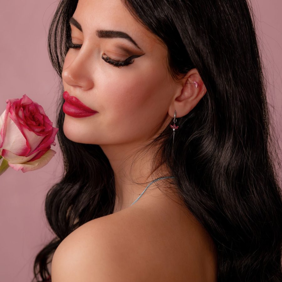 Frau mit langen dunklen Haaren hält eine rosa Rose und trägt einen silbernen Ohrring mit rotem Kristall auf rosa Hintergrund