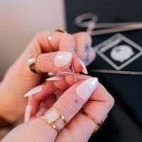 Hände mit langen, weißen Nägeln halten eine Piercingnadel vor einem schwarzen Hintergrund