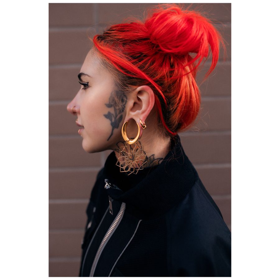 Frau mit roten Haaren trägt goldene Kaleidoscope Hoops vor einer Backsteinwand