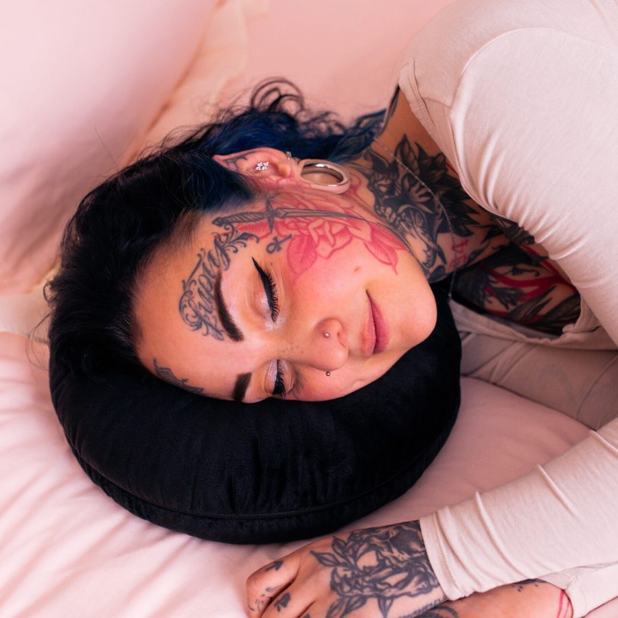 Person mit Tattoos liegt auf einem schwarzen Piercingkissen auf rosa Bettwäsche