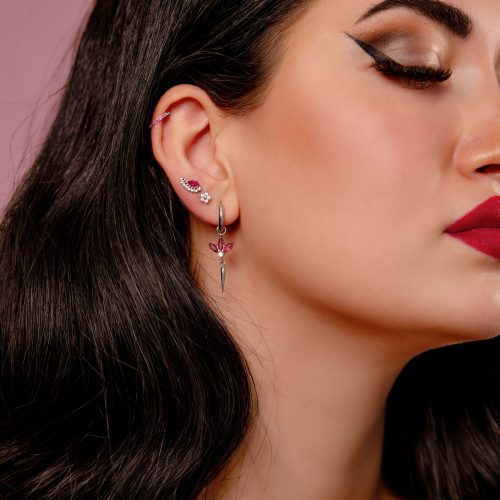 Nahaufnahme eines Ohrs mit mehreren Piercings, darunter ein silberner Clicker mit roten Kristallen, auf rosa Hintergrund.