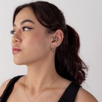 Frau mit dunklem Haar trägt schwarzen Septum Clicker und mehrere Ohrringe in verschiedenen Formen vor einem weißen Hintergrund.