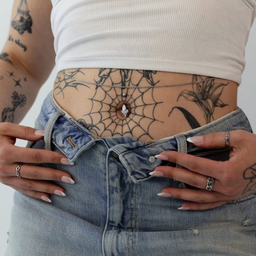 Person mit Tattoos trägt ein silbernes Bauchnabelpiercing mit Kristall auf Jeans und weißem Top