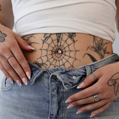 Nahaufnahme eines Bauchnabels mit silbernem Bananabell und Kristall, umgeben von Tattoos. Hände mit lackierten Nägeln halten die Jeans.