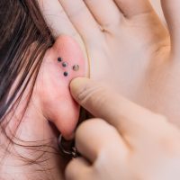 Nahaufnahme eines Ohrs mit drei schwarzen Labret Piercings und einem Finger, der die Piercings berührt.