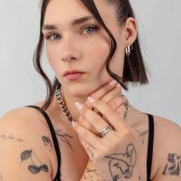 Frau mit kurzen Haaren trägt silberne Ohrringe, Halskette und Ringe. Tätowierungen sichtbar, grauer Hintergrund.