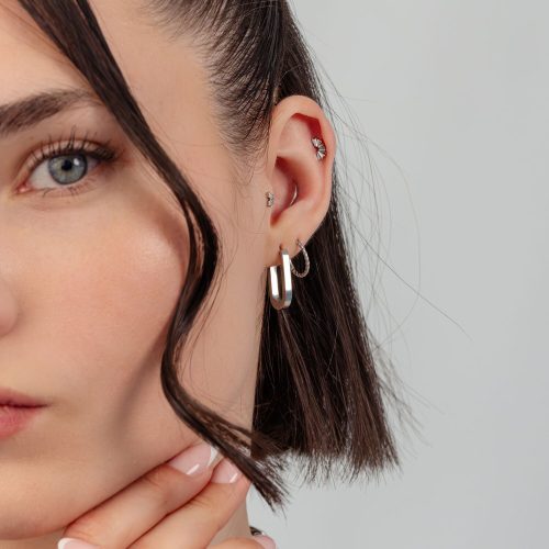 Nahaufnahme eines Ohres mit silbernen Creolen und mehreren Ohrpiercings vor grauem Hintergrund.