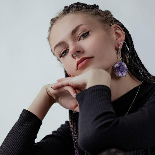 Junge Frau mit Zöpfen, Amethyst-Ohrring, Nasenpiercing, schwarzem Pullover vor grauem Hintergrund