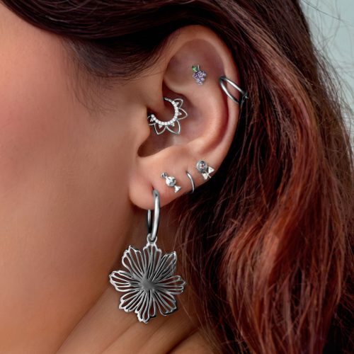 Nahaufnahme eines Ohrs mit mehreren silbernen Piercings, darunter ein großer Blumenanhänger, auf hellem Hintergrund