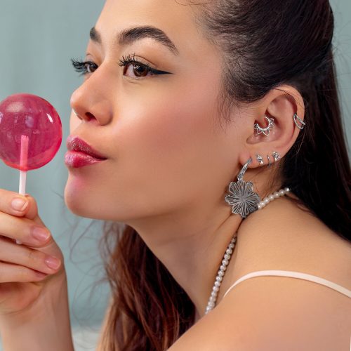 Frau mit mehreren silbernen Ohrpiercings und einem rosa Lollipop, seitlich