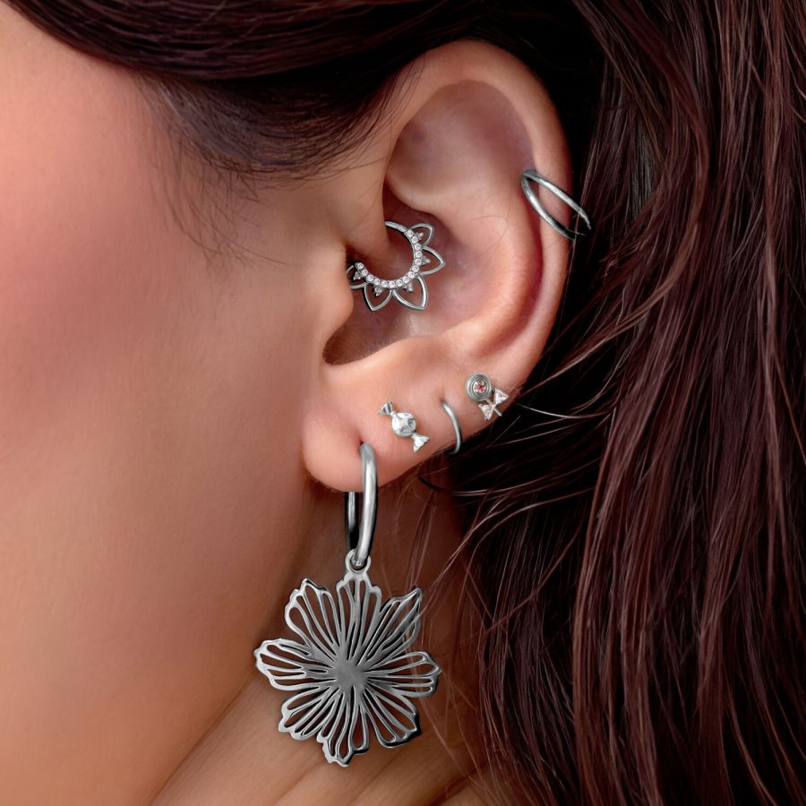 Nahaufnahme eines Ohres mit mehreren silbernen Piercings, darunter ein großer Blumenanhänger und ein filigraner Ring.