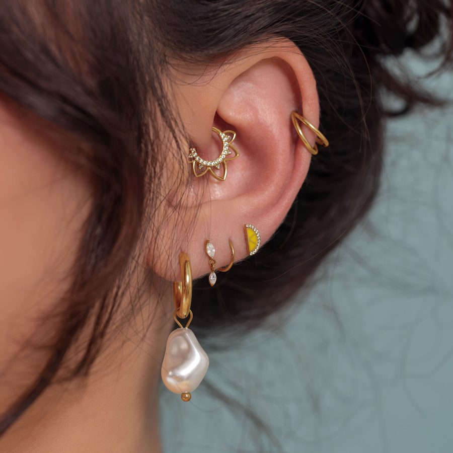 Nahaufnahme eines Ohrs mit mehreren goldenen Piercings, darunter ein Perlenanhänger, auf einem hellblauen Hintergrund