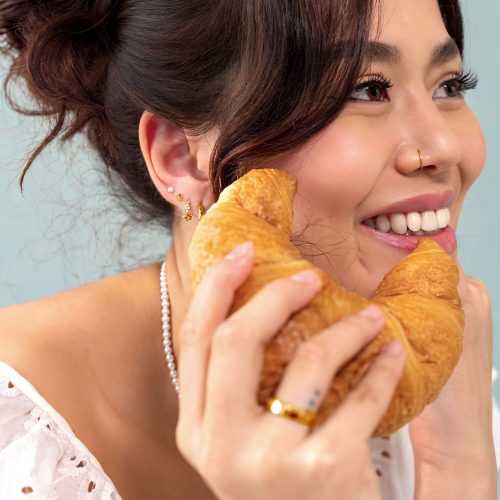 Frau hält Croissant an Gesicht, trägt goldene Piercings, lächelt