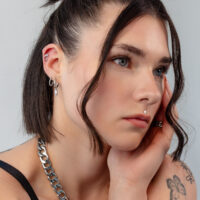 Nahaufnahme eines Gesichts mit mehreren silbernen Ohrpiercings und Kristalldetails auf grauem Hintergrund