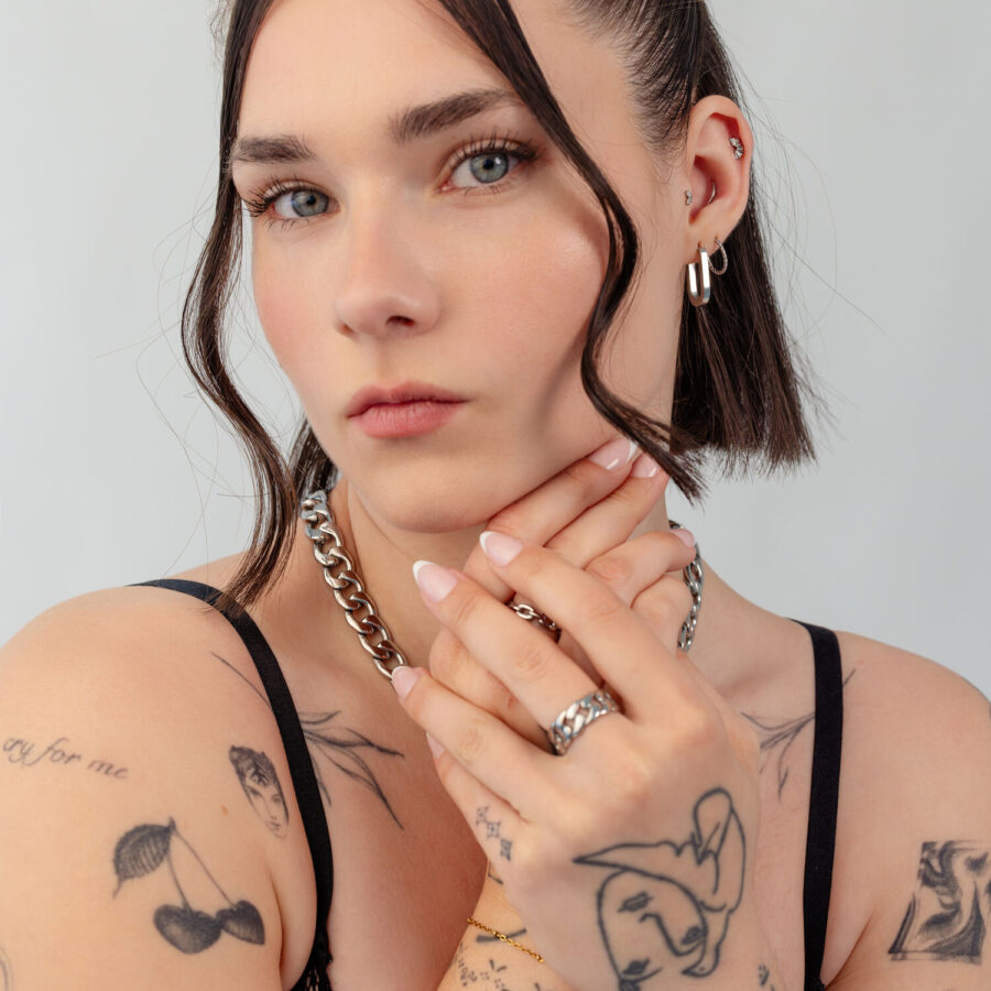Frau mit kurzen Haaren trägt silberne Kette und Ohrringe, Tattoos sichtbar