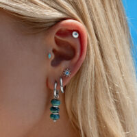 Ohr mit mehreren Piercings, inklusive türkisfarbener Kristallaufsatz und silbernen Creolen, vor blauem Himmel