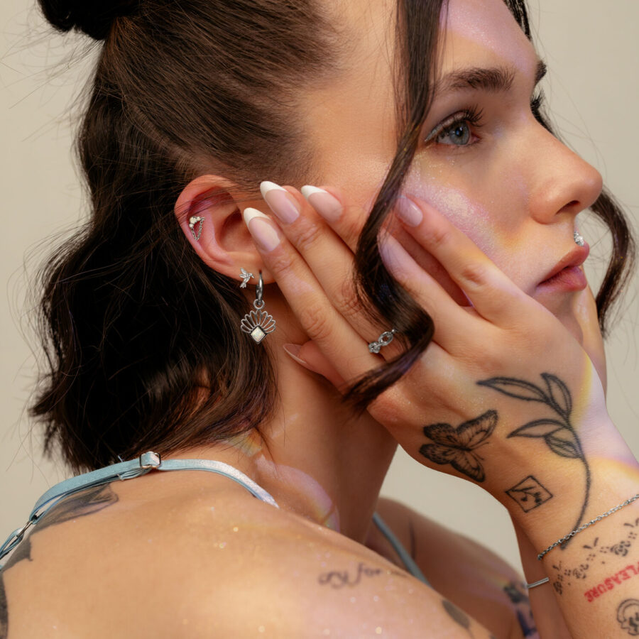 Junge Frau mit getöntem Licht auf dem Gesicht, trägt einen silbernen Ohrstecker mit Schmetterlingsanhänger und hat Tattoos auf den Händen.