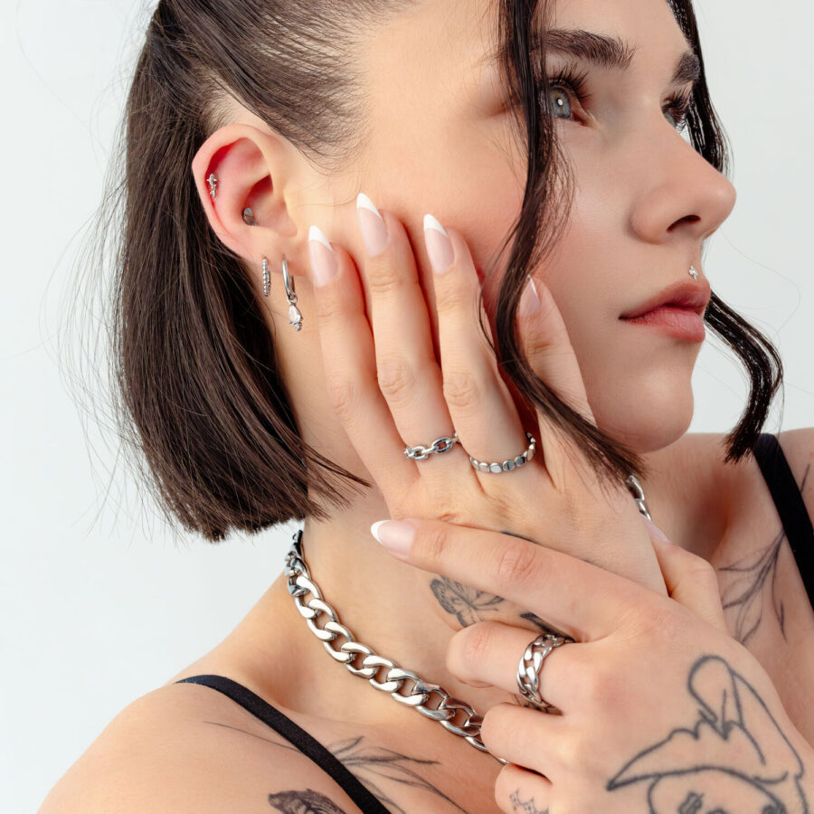 Frau mit Tattoos trägt silberne Ringe, Ohrringe und Kette, Nahaufnahme auf hellem Hintergrund