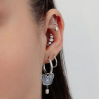 Nahaufnahme eines Ohres mit mehreren silbernen Piercings, Kristallen und einem Schmetterlingsanhänger auf einem weißen Hintergrund.
