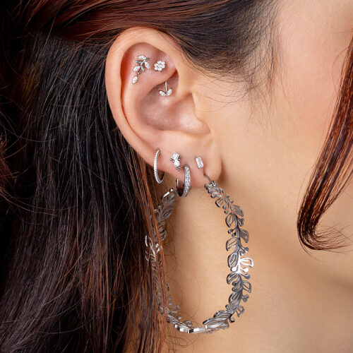 Ohr mit mehreren Piercings, darunter silberne Creolen und Kristallaufsätze, braunes Haar im Hintergrund