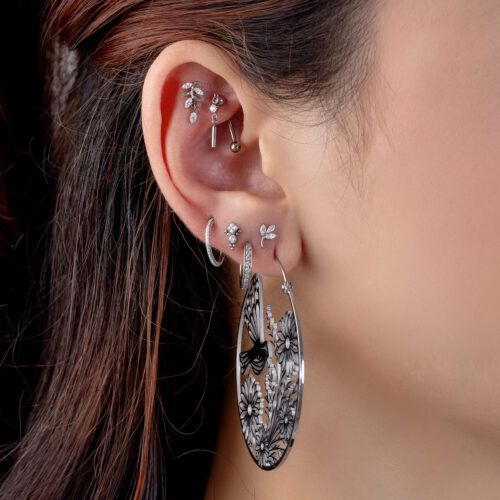Ohr mit mehreren silbernen Piercings, darunter ein großer runder Ohrring mit floralem Muster und klare Kristalle.